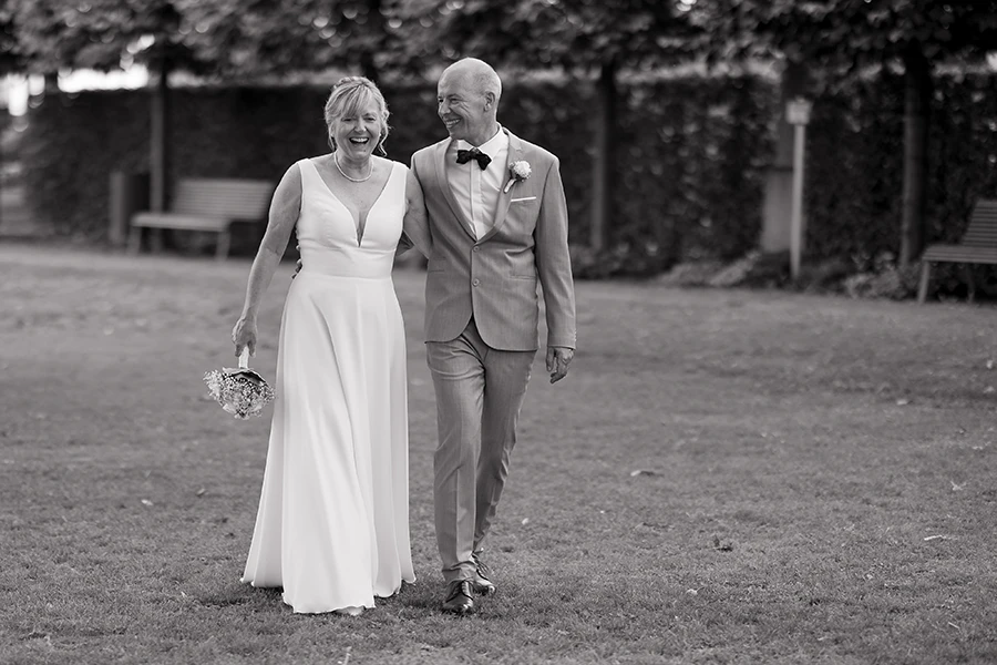 Schwarz-weiß Foto wie das Brautpaar Arm in Arm lachend auf die Kamera zuläuft.